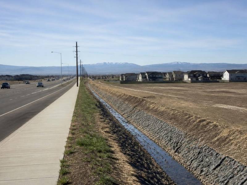 Denver roadway beside an undeveloped site with a sidewalk, drainage channel, nearby homes, and mountains in the distance.